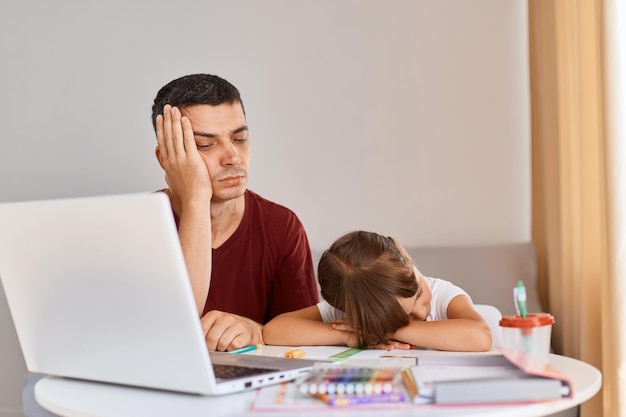 Tiro interior de pai adulto jovem sonolento cansado sentado com filha colegial na mesa família posando com os olhos fechados sendo exausto para fazer tarefas difíceis