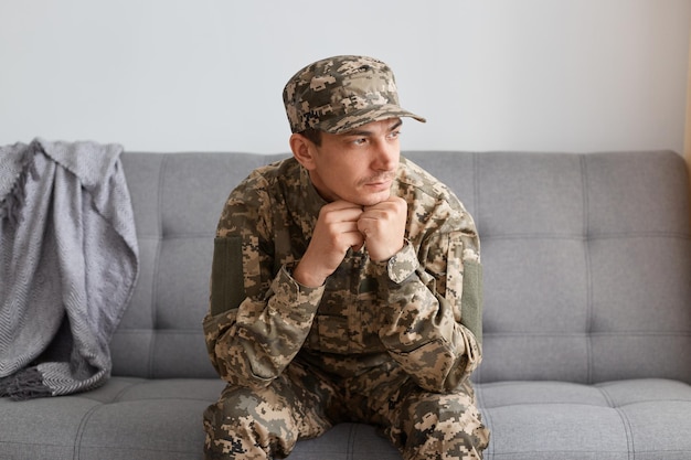 Tiro interior de soldado triste em uniforme de camuflagem sentado em um sofá e segurando os punhos sob o queixo olhando para longe com expressão facial pensativa voltando para casa da guerra