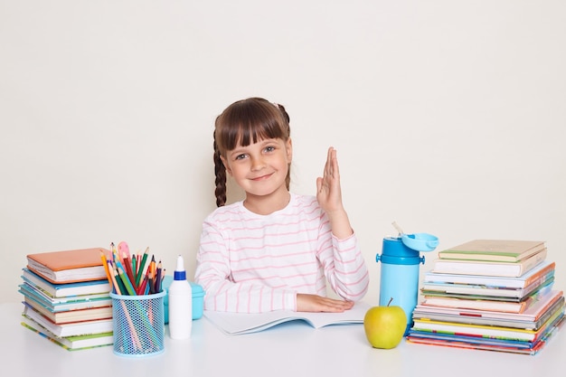 Tiro interior de sorridente garotinha encantada com cabelos escuros e tranças, sentado à mesa cercada de livros e criança de braço levantado quer responder durante a aula