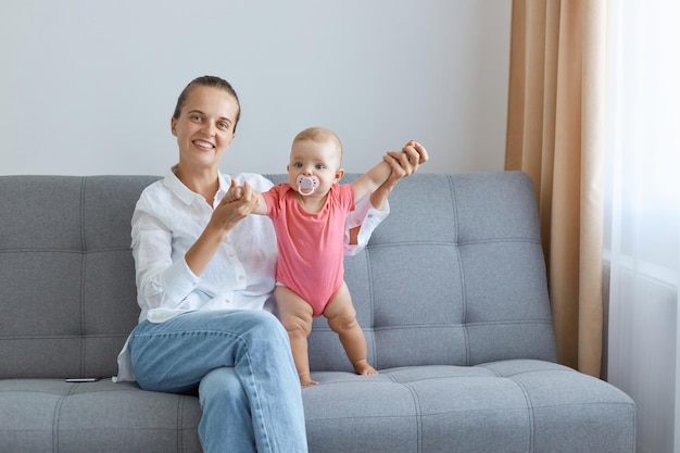 Tiro interior de sorridente mulher satisfeita vestindo camisa branca e jeans, sentado no sofá com a filha filha mãe segurando as mãos da criança olhando para a câmera expressando felicidade