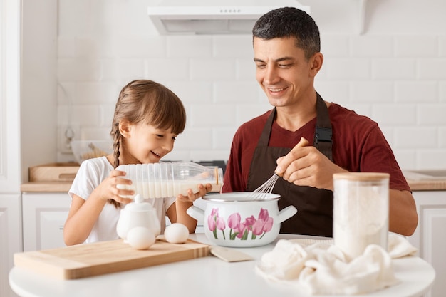 Tiro interior de sorridente pai positivo sentado à mesa com a filha e cozinhando juntos na cozinha menina derramando leite na panela e sorrindo vai fazer panquecas