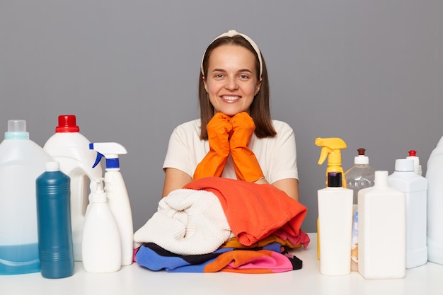 Tiro interior do zelador de mulher sorridente mantém as mãos sob o queixo parece feliz usando bandana e camiseta casual usando detergentes e esponjas para lavanderia isolada sobre fundo cinza