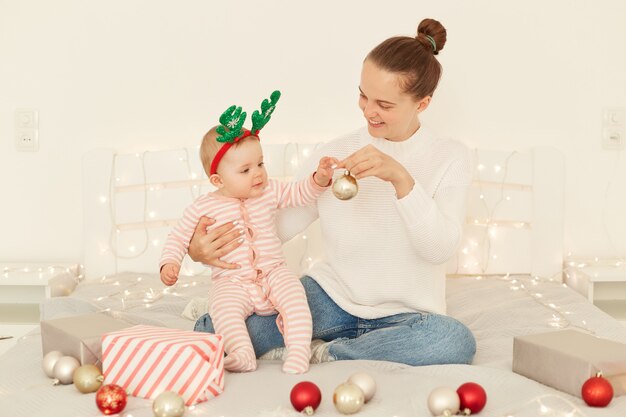 Tiro interno da bela mãe jovem adulta sentada na cama branca, segurando um bebê nos braços, sorrindo, comemorando o Natal, brincando com enfeites de ano novo.