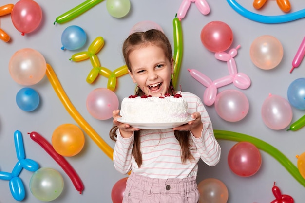 Tiro interno da menina feliz aniversário com tranças vestindo roupas casuais posando isolado sobre fundo cinza com balões segurando mordendo seu bolo com cerejas