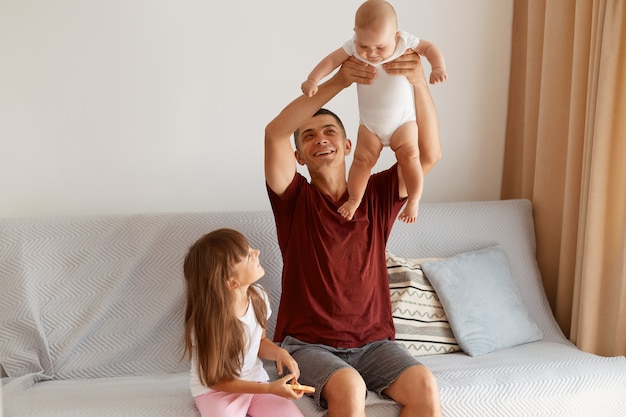 Tiro interno da morena jovem adulto caucasiano homem vestindo camiseta estilo casual cor de vinho, posando na sala de estar em casa, sentado no sofá com seus dois filhos, vomitando bebê infantil.