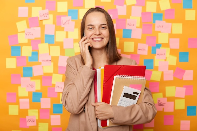 Tiro interno da mulher atraente sorridente amigável na jaqueta segurando organizador e pasta de papel falando no celular com expressão feliz em pé contra a parede amarela com adesivos multicoloridos