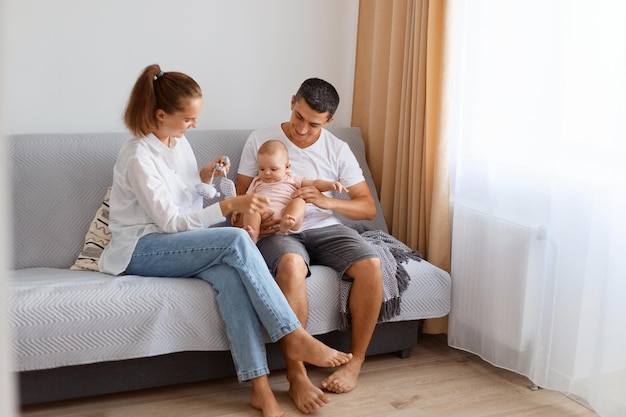 Tiro interno de casal jovem feliz, vestindo roupas de estilo casual, sentado no sofá, na sala de estar iluminada com um bebê infantil, pais brincando com sua filha, paternidade e infância.