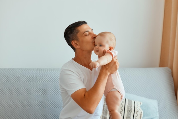 Tiro interno de cuidar de homem bonito, vestindo camiseta branca, sentado no sofá e beijando sua encantadora filha pequena, passando o fim de semana com a criança.