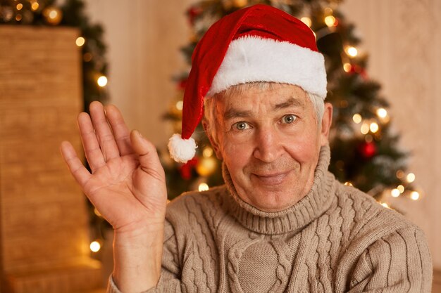 Tiro interno de homem maduro otimista positivo vestindo suéter quente e chapéu de papai noel, olhando para a câmera e acenando com a mão, posando na festiva sala de estar em casa perto da árvore de natal.