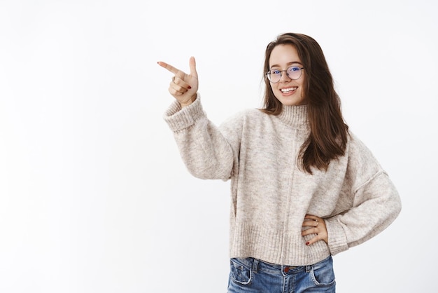 Tiro interno de mulher atraente despreocupada feliz de óculos e suéter segurando a mão na cintura sorrindo e rindo como apontando para o canto superior esquerdo encantado e determinado na escolha sobre a parede cinza