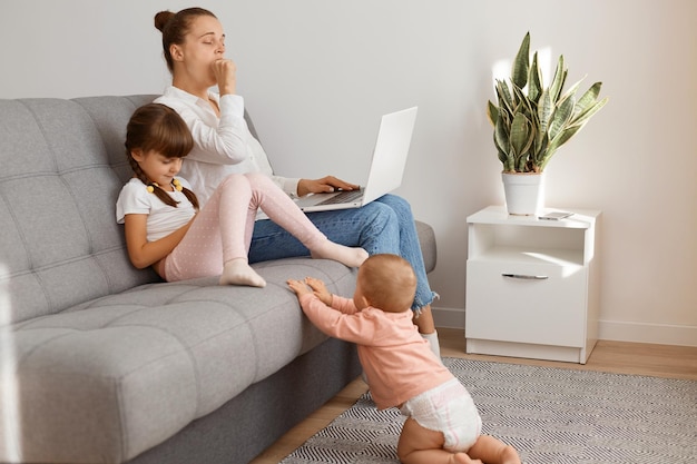 Tiro interno de mulher cansada com sono, vestindo camiseta e calça jeans, sentada no sofá com seus filhos, bocejando, exausta por trabalhar longas horas na frente do laptop, cobrindo a boca com a mão.