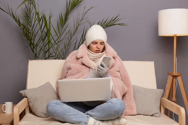 Tiro interno de mulher chocada usando cachecol de luvas de boné e casaco sentado no sofá com o laptop e usando o telefone tendo problemas com seu trabalho digitando no celular