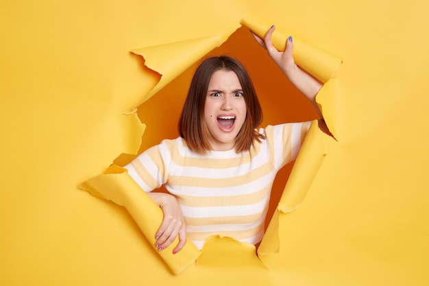 Tiro interno de mulher de cabelos escuros agressivo com raiva vestindo camisa listrada olhando pelo buraco no papel amarelo expressando raiva gritando estar de mau humor