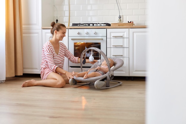 Tiro interno de mulher de cabelos escuros, vestindo camisa listrada estilo casual e curta sentado no chão perto do forno com filha infantil na cadeira de balanço, preparando bolo ou torta, esperando por pastelaria.