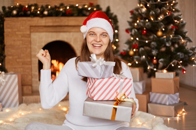 Tiro interno de mulher feliz e animada com suéter branco e chapéu de Papai Noel, estando na sala festiva com lareira e árvore de Natal, sentado com uma pilha de presentes de ano novo.