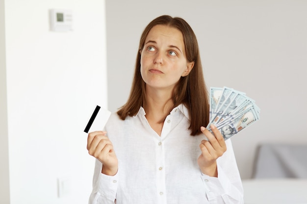 Tiro interno de mulher pensativa, vestindo uma camisa branca estilo casual, posando na sala de luz em casa, olhando para longe, segurando dinheiro e cartão de crédito nas mãos, pensa como gastar dinheiro.