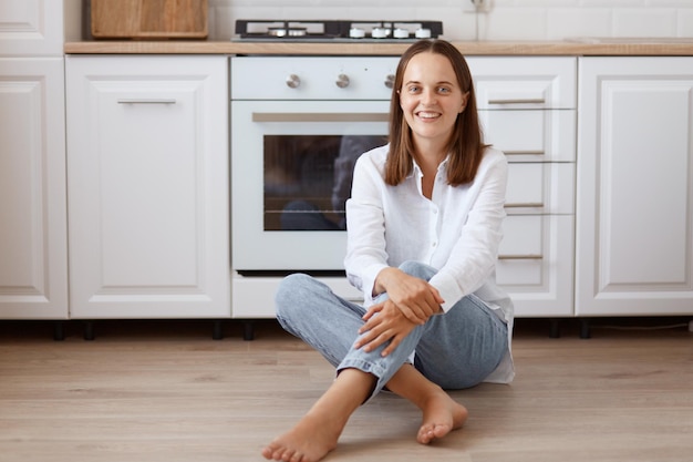 Tiro interno de mulher sorridente com cabelo escuro, sentada no chão da cozinha e olhando para a câmera com uma expressão positiva, vestindo jeans e camisa branca.