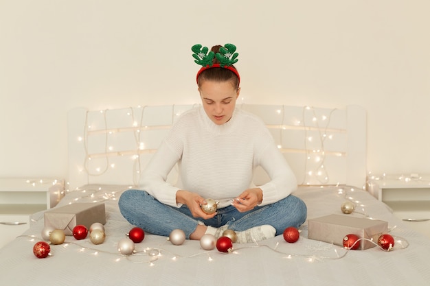 Tiro interno de mulher vestindo jeans e suéter branco casual, localização com as pernas cruzadas na cama, segurando bugiganga de Natal nas mãos, tendo expressão facial negativa.