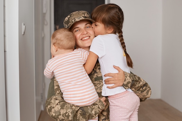 Tiro interno de soldado patriótico jovem adulto vestindo uniforme de camuflagem, voltando para casa após o serviço militar e abraçando seus filhos com amor.