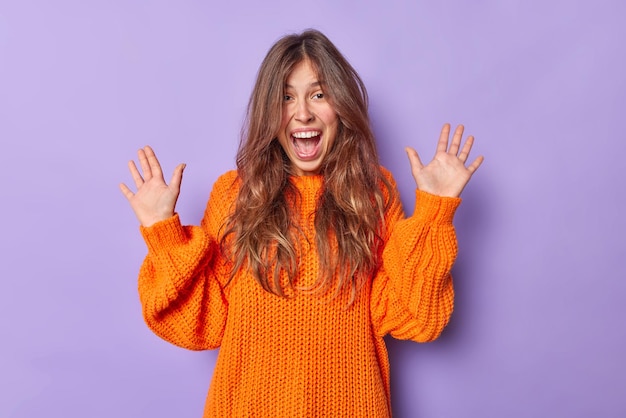 Tiro interno de uma jovem emocional exclama em voz alta mantém as palmas das mãos levantadas e reage alegremente às notícias incríveis, vestida com um suéter de malha solto isolado sobre o fundo roxo. conceito de felicidade.