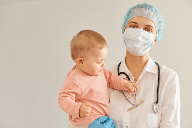 Tiro interno do médico pediatra usando boné médico, máscara cirúrgica, vestido e luvas de borracha, olhando para a câmera com um sorriso, em pé com a menina da criança nos braços.