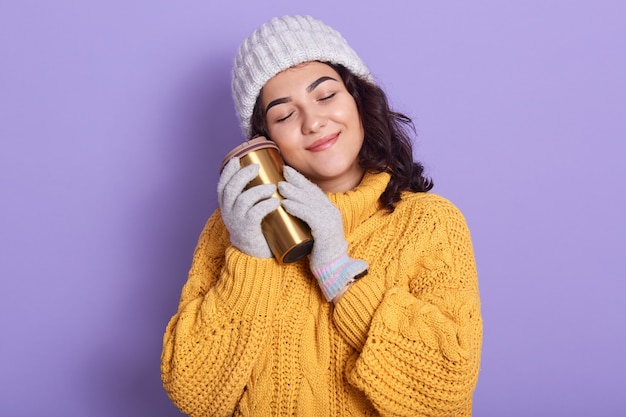 Tiro interno horizontal do concurso pacífica morena jovem, fechando os olhos, sorrindo, segurando o thermomug, colocando-o perto do rosto, usando chapéu branco, luvas e blusa amarela brilhante.