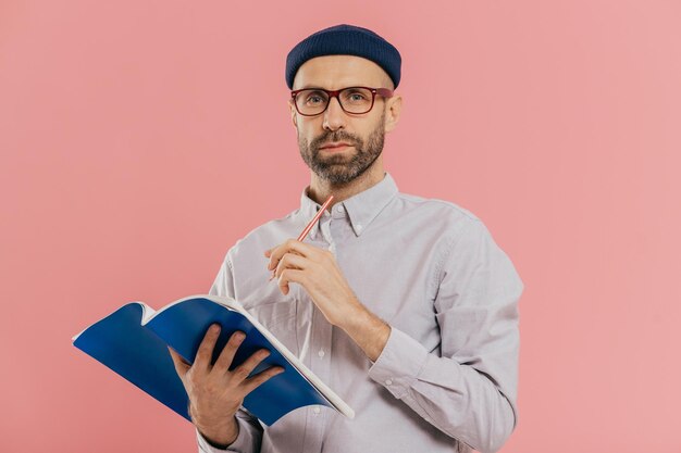 Tiro isolado de homem jovem atraente segura livro faz anotações no organizador vestido formalmente Estudante inteligente se prepara para exame ou faz tarefa de casa isolada sobre parede rosa do estúdio