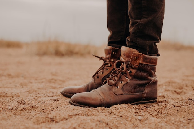 Tiro lateral do homem em sapatos marrons desgrenhados na superfície leiga Par de botas na areia Macho tem caminhada ao ar livre em calçados velhos