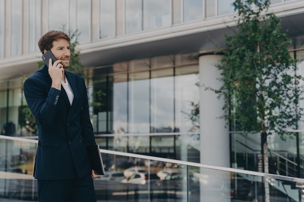 Tiro lateral de llamadas de hombre barbudo a través de un teléfono inteligente vestido con traje formal negro posa al aire libre