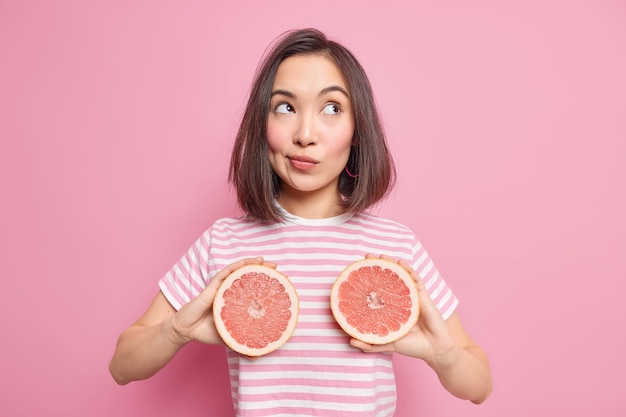 Tiro de media longitud de una chica milenaria pensativa con apariencia oriental sostiene dos mitades de toronja fresca come cítricos que contienen vitamina C tiene una alimentación saludable. Vitaminas para la belleza y el cuidado de la piel.