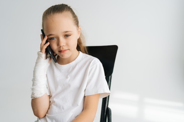 Tiro médio da menina loira com braço quebrado envolto em bandagem de gesso falando smartphone olhando para câmera sentado cadeira no fundo branco