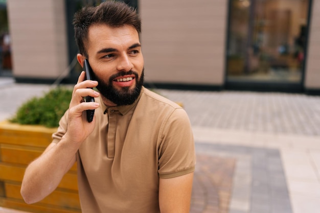 Tiro médio de jovem alegre falando no smartphone sentado no banco na rua da cidade no fundo do edifício homem barbudo conversando