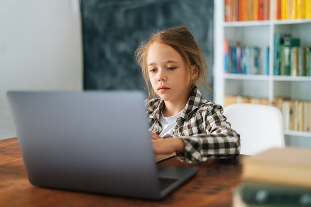Tiro médio de menina da escola primária de criança séria sentada à mesa com o laptop e o livro de papel na acolhedora sala das crianças, olhando para a tela. linda aluna do ensino fundamental e-learning on-line usando o computador.