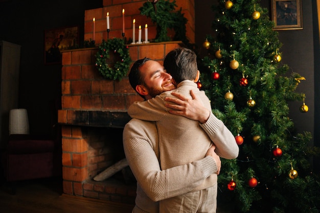 Tiro médio do filho criança feliz abraçando o pai jovem e alegre perto da árvore de Natal decorada com bolas e lareira aconchegante na sala escura
