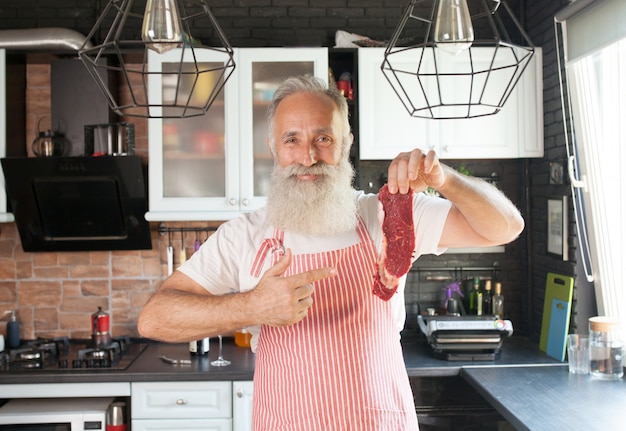 Tiro medio del hombre barbudo sonriente que sostiene un pedazo de carne cruda. Sosteniendo la carne fresca aislada en la cocina. De pie en la cocina.