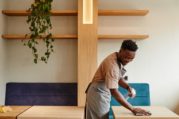 Tiro medio largo de joven afroamericano vistiendo delantal trabajando en un pequeño café preparando mesas ...