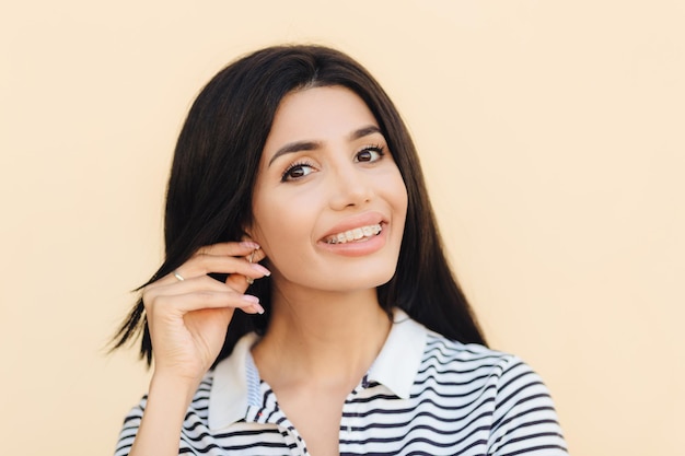 Tiro na cabeça de linda mulher bonita tem sorriso largo cabelo escuro mantém a mão na orelha mostra dentes brancos com aparelho olha diretamente para a câmera tem expressão feliz isolada sobre o fundo do estúdio