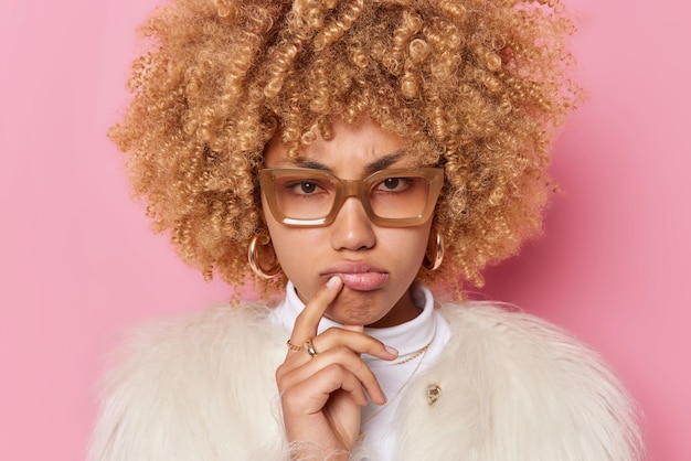 Tiro na cabeça de mulher de cabelo encaracolado descontente franze os lábios parece sério ouve algo atentamente usa óculos e casaco de pele isolado sobre fundo rosa se sente envergonhado sendo ofendido