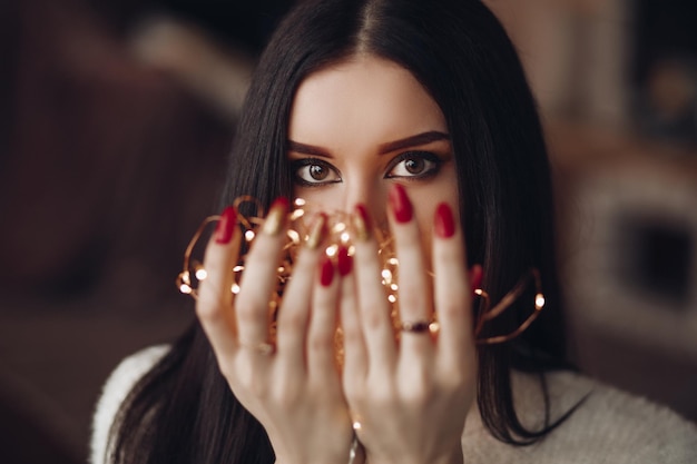 Foto tiro na cabeça de mulher de cabelos escuros com maquiagem de noite brilhante segurando um monte de luzes nas mãos com unhas vermelhas