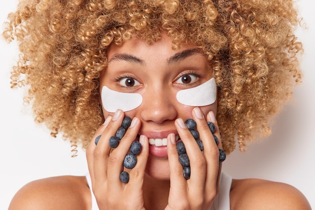 Tiro na cabeça de uma linda mulher alegre sorri com prazer tem mirtilos entre os dedos aplica almofadas de beleza sob os olhos para remover o inchaço fica ombros nus isolados sobre fundo branco