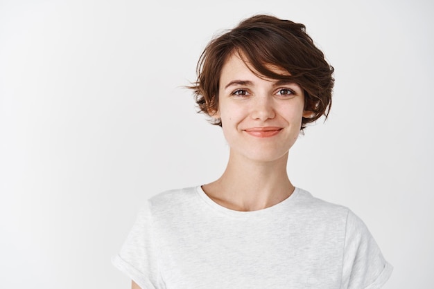 Tiro na cabeça de uma linda mulher caucasiana com corte de cabelo curto sorrindo e parecendo confiante em camiseta em fundo branco