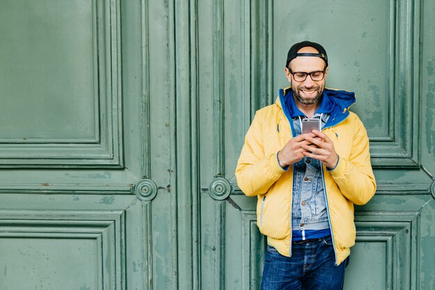 Tiro na cabeça do homem elegante, com barba e testa larga em anoraque amarelo, jeans e boné em grandes óculos, segurando o telefone celular