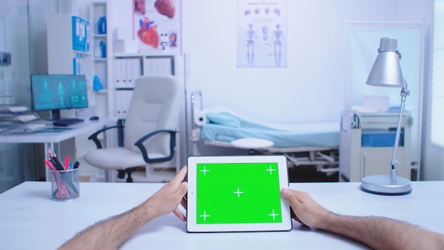 Tiro pov de asistente sosteniendo tablet pc con pantalla verde en el gabinete del hospital y médico con bata blanca trabajando en computadora. Medic con tableta con clave de croma en exhibición en la clínica médica.