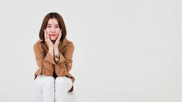 Tiro del recorte del estudio del modelo femenino asiático pensativo joven del pelo bastante corto en los zapatos casuales de la chaqueta de la capa marrón larga que se sientan piernas cruzadas pensando decidir resolver la idea en la silla del cuadro en el fondo blanco.