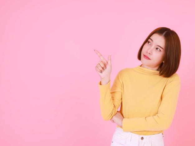 Tiro del recorte del estudio del retrato del modelo femenino asiático del pelo bastante corto joven en la sonrisa amarilla del soporte de la camisa de manga larga para mirar a la cámara y que señala el dedo sobre fondo rosa.