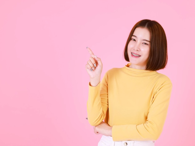 Tiro del recorte del estudio del retrato del modelo femenino asiático del pelo bastante corto joven en la sonrisa amarilla del soporte de la camisa de manga larga para mirar a la cámara y que señala el dedo sobre fondo rosa.