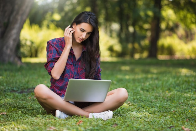 Tiro sincero de jovem morena caucasiana com um sorriso freelancer vestido casualmente sentado no gramado em frente ao notebook trabalhando em seu laptop fazendo compras lendo artigos estudando