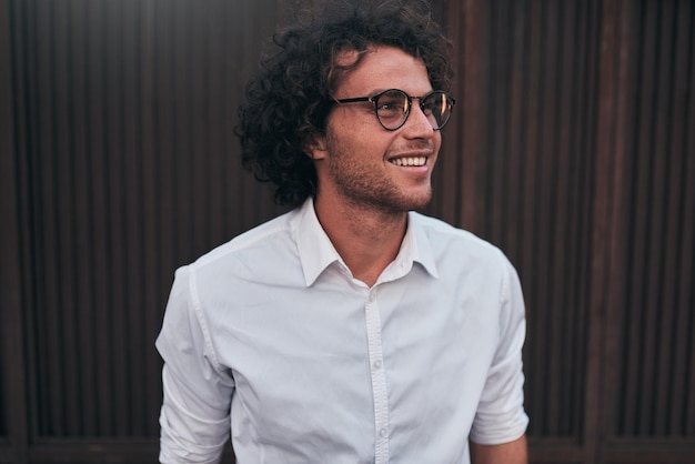 Tiro urbano do jovem empresário bonito com óculos sorrindo posando ao ar livre Estudante do sexo masculino posando contra a parede marrom Cara inteligente na camisa branca usa óculos com cabelos cacheados andando na rua