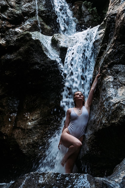 Tiro vertical de una atractiva mujer sexy en traje de baño posando bajo una cascada que fluye sobre rocas