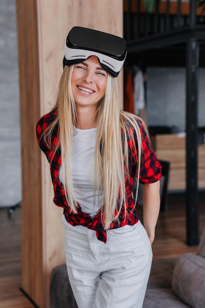 Tiro vertical de atraente jovem loira caucasiana vestida com camisa xadrez e calça branca com óculos vr na cabeça em pé em casa sorri muito feliz em jogar usando fone de ouvido de realidade virtual